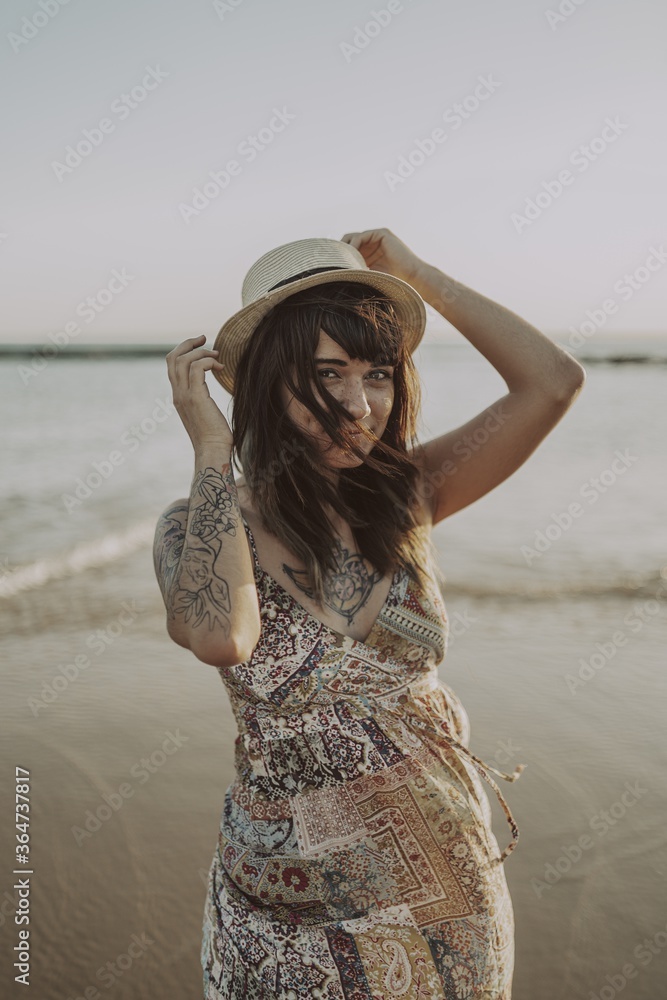 Wall mural young female with tattoos wearing a dress and straw hat on blurred ocean background