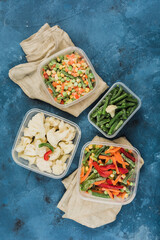 Frozen vegetables: a mix of vegetables, string beans and cauliflower in different plastic containers for freezing with napkins on a blue background.