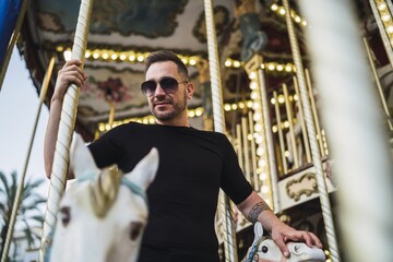 Young Caucasian male spending fun time in an attraction park with a carousel with horses at daytime