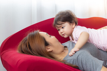 Infant baby or adorable little mix race baby look at her asian mother and lying down on her mother chest with smile face. Lovely baby stay with single mother together on red sofa in living room