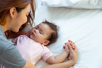 Asian mother lull kid, adopt little girl to be her protege or daughter Single mother admire with love, fondness of caucasian infant girl while sleeping on bed. Lovely toddler baby holding mom finger