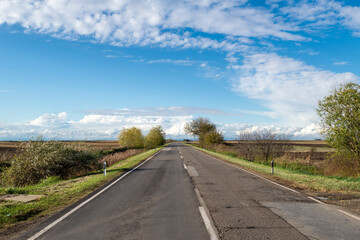 Cloudy country repaired highway/road in bad condition and blue cloudy sky. Green and yellow/orange fields, sunny landscape.