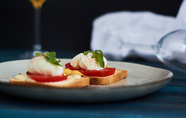 Large gray ceramic plate with toast and poached eggs, greens, tomatoes, sauce. Tasty breakfast. Glasses of wine.