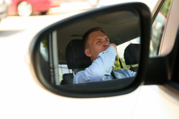 Tired young man yawning in his auto, view through car side mirror