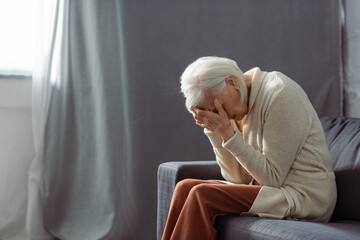 depressed senior woman sitting on sofa with bowed had and obscuring face with hands