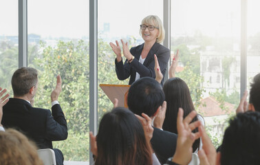Senior female business speaker stand clapping hands on podium with the audience in seminar event,...