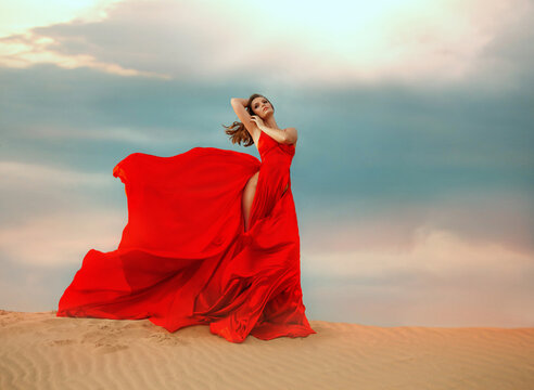young sexy woman glamour queen stands. Beautiful desert sand dune dramatic sky clouds. Red long silk elegant satin dress, slit up leg skirt, fluttering fly in wind motion. Girl fashion model posing
