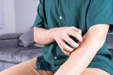 Young man scratching his arm while sitting on sofa at home. Healthcare medical or daily life concept.