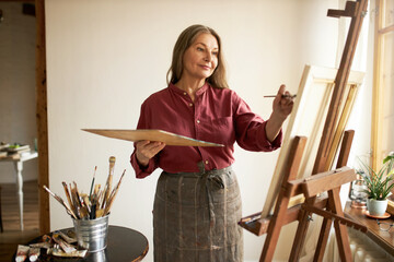 Beautiful elderly gray haired female artist working in her studio holding palette, applying acrylic paint on canvas using paintbrush, smiling. Inspiration, creative skills and art concept