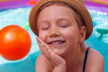 Young girl playing in colorful rainbow inflatable swimming pool with orange ball. Happy child in straw hat smiling in clear blue water. Summer active lifestyle, children swim fun leisure hot weather.