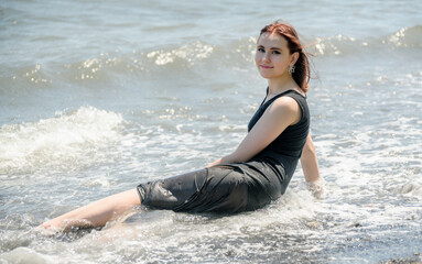 Fototapeta na wymiar Young woman in black dress sitting on sand and enjoys sea.