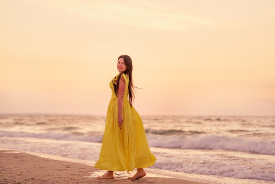 A girl in a long dress with long black hair walks at dawn along a deserted beach along the line of the surf. 