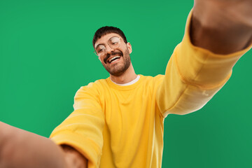 people concept - happy smiling young man in glasses and yellow sweatshirt making selfie over...