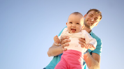 Father holds a small child in his arms on a background of a summer blue sky. Cute baby is smiling. Happy family outdoors. Right space for text.