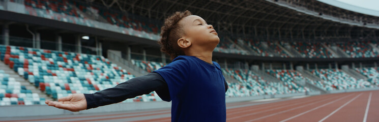 Cute little black kid boy spreading his hands on an empty stadium, dreaming of becoming...