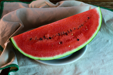 Slice of watermelon on white plate, green-bordered cotton tablecloth and wooden table.