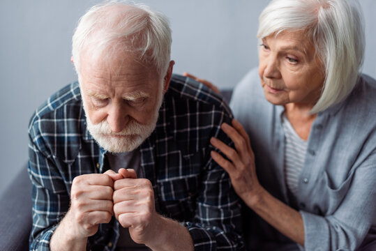 worried senior woman touching husband suffering from dementia and sitting with clenched fists