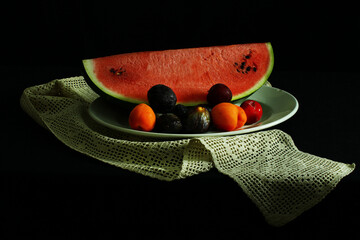 Still life. White plate with watermelon, figs, apricots and plums. Crochet doily and black...