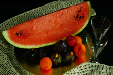 Still life. White plate with watermelon, and various fruit. Crystal glasses to the side. Crochet...