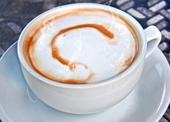 White cup and saucer with coffee latte has the letter C dribbled in the foam for a type of coffee art.  Piping hot steam is coming off the beverage.