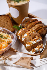 Disposable tableware, soup in a paper cup, bread, salad and baked potatoes with cottage cheese and nuts on a wooden tray on a white background.