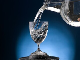 Jug is pourung water into a stem glass. Close-up on blue background