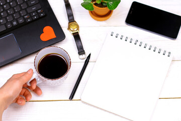 Laptop with red paper heart, notebook, pan, phone, hand clock, green leaves, womanâ€™s hand with coffee cup on old white wooden background. Romantic Work Table concept.