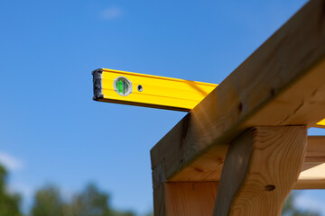 A wooden beam made of fresh timber with yellow carpentry tools - a water level for accurate marking on a summer day against the sky and green tree tops.