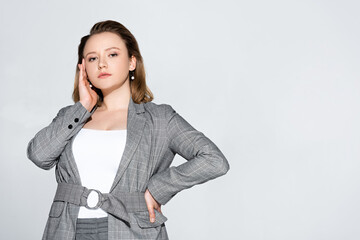 attractive, stylish plus size girl touching face while holding hand on hip isolated on grey