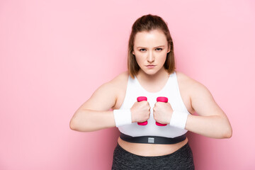 serious overweight girl training with dumbbells while looking at camera on pink
