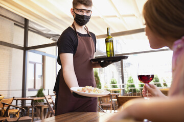 The waiter works in a restaurant in a medical mask, gloves during coronavirus pandemic. Representing new normal of service and safety. Putting the order, meals and drink, wine. Taking care of clients.