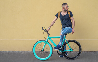 portrait of fashion young cyclist, riding a minimalist bike with yellow wall on background, going from home to work, an ecological and sustainable city transport concept