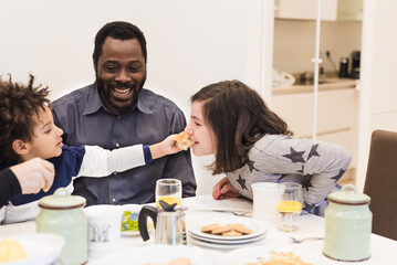 Happy black father with young children having breakfast, mixed-race family having fun in the kitchen, the little boy plays and feeds his sister and daddy looks them and smiles