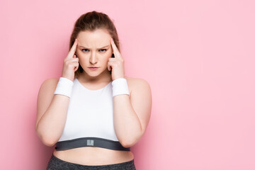 serious, thoughtful overweight girl touching head while looking at camera on pink
