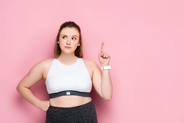 thoughtful overweight girl showing idea sign while holding hand on hip on pink