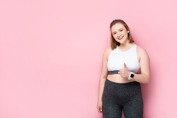 happy overweight girl showing thumb up while smiling at camera on pink