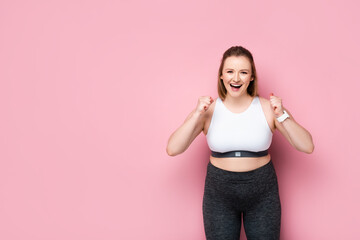 excited overweight girl in sportswear showing winner gesture on pink