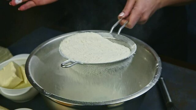 slow motion closeup woman hands sifting flour by metal sieve into empty steel bowl