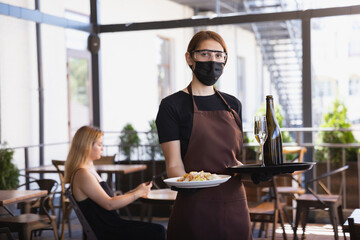 The waitress works in a restaurant in a medical mask, gloves during coronavirus pandemic. Representing new normal of service and safety. Putting the order, meals, drinks,. taking care of clients.