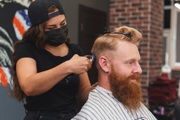 Female barber in mask cuts a man hairs with hair clipper. hairstyle during social distancing