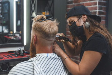 Female barber in mask cuts a man hairs with hair clipper. Hairstyle during social distancing