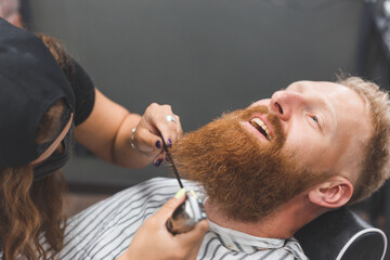 A man at a barbershop. Woman barber clipping beard and mustache. Barber woman in mask.