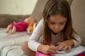 White child girl studying at home, near toys, playing teaching dolls, holding notebook sheet