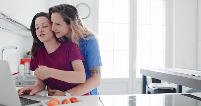 Portrait of loving lesbian couple spending good time together in the kitchen, kissing and looking at their notebook. LGBT concept.