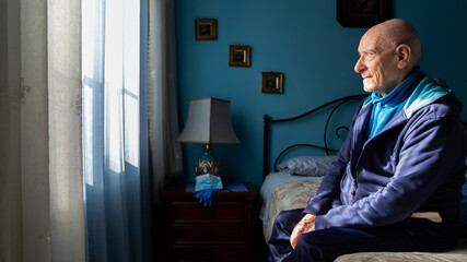 Covid-19 Profile view of an old man sitting on his bed looking sad at the window