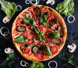 Pizza with mushrooms, onions, and tomatoes lies on a dark table with sprinkled salt and pepper