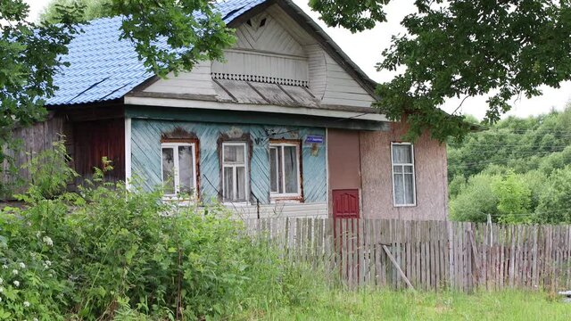 An old rustic wooden house stands behind a low fence. It is painted in blue. Roof tiles. Around the green bushes, grass and trees. 4K.