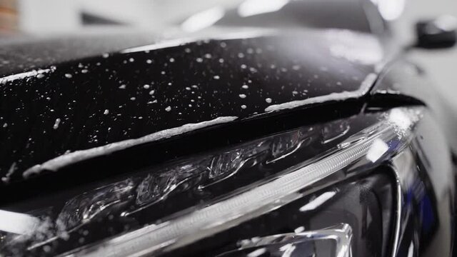 Slow motion pan across the front of a black car as sudsy spray wets the hood of the car.