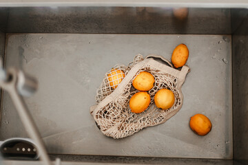 Yellow lemons are in net cotton shopping bag in sink. Washing fruits before using them. Local farm produce. Fresh seasonal food. Bright color. Zero waste, organic, healthy. From farm to table. Citrus
