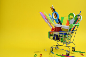Colored pencils in a small shopping cart on a yellow background with space for text. Back to school, office supplies.
